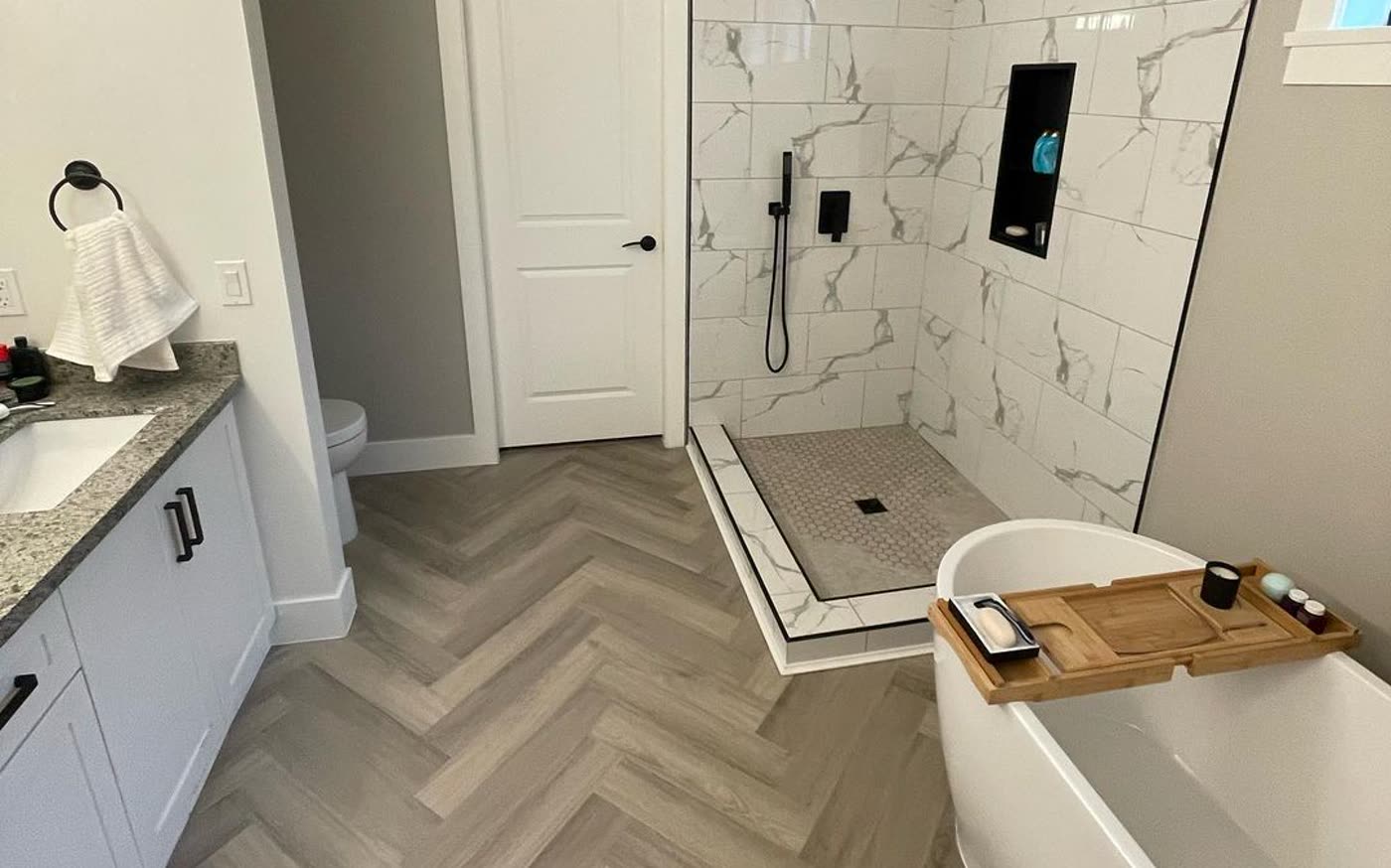 A renovated ensuite with a freestanding tub, walk-in shower tiled in marble-look porcelain and a herringbone floor.