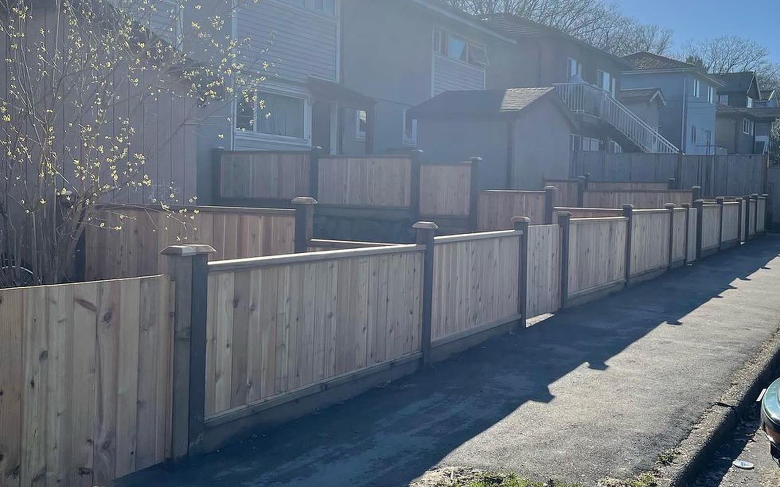 A new cedar fence with capped posts running along a shared walkway at a multi-unit residential property, with a second run enclosing unit patios across the lawn.