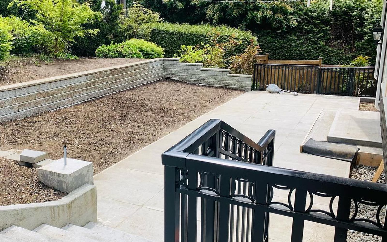 A terraced back garden with stepped concrete block retaining walls, a poured concrete patio and black metal railings, backed by mature hedging.
