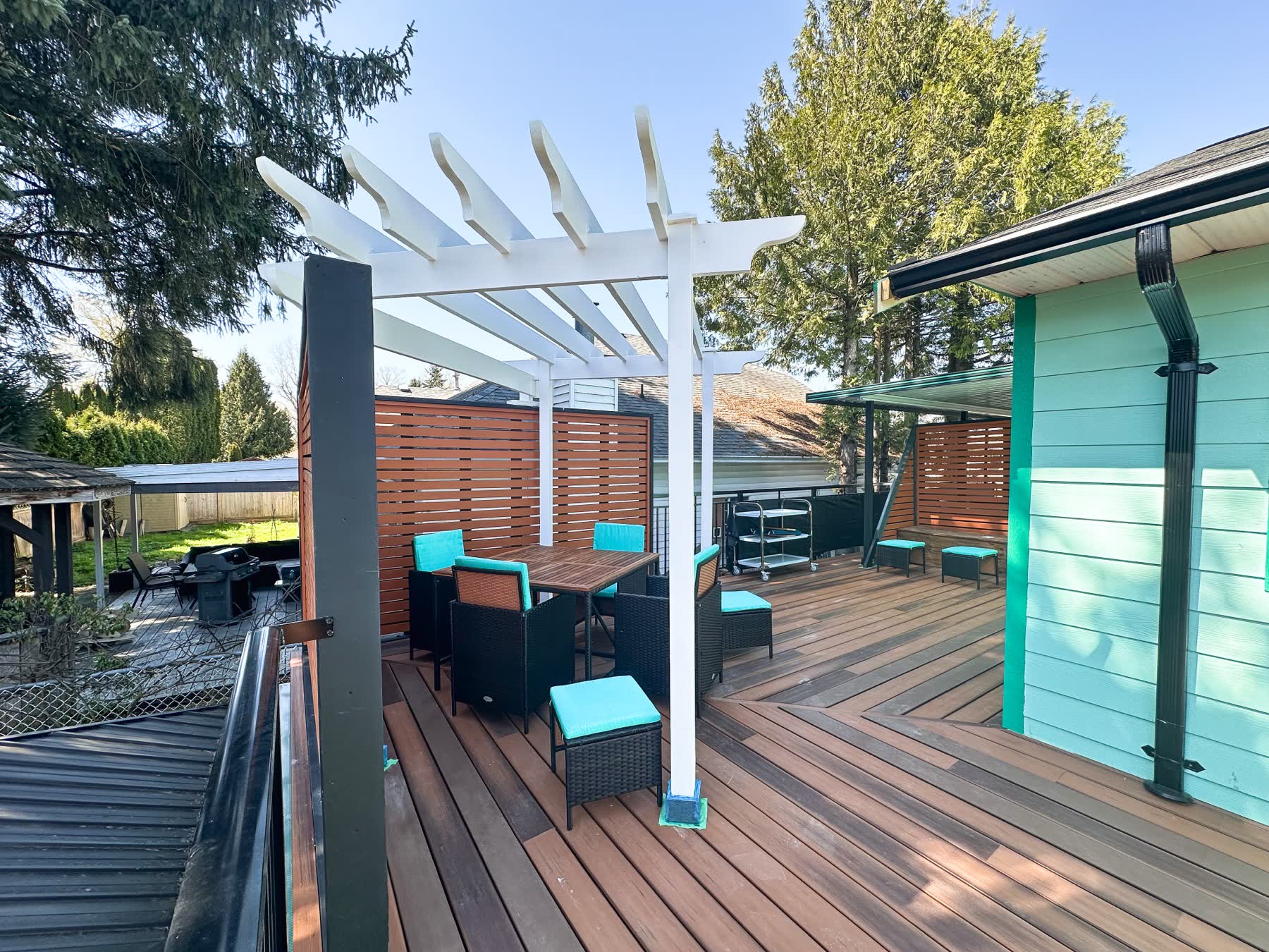 Rear deck in Maple Ridge with a white pergola, slatted privacy screens and patterned hardwood decking beside a teal-painted house.
