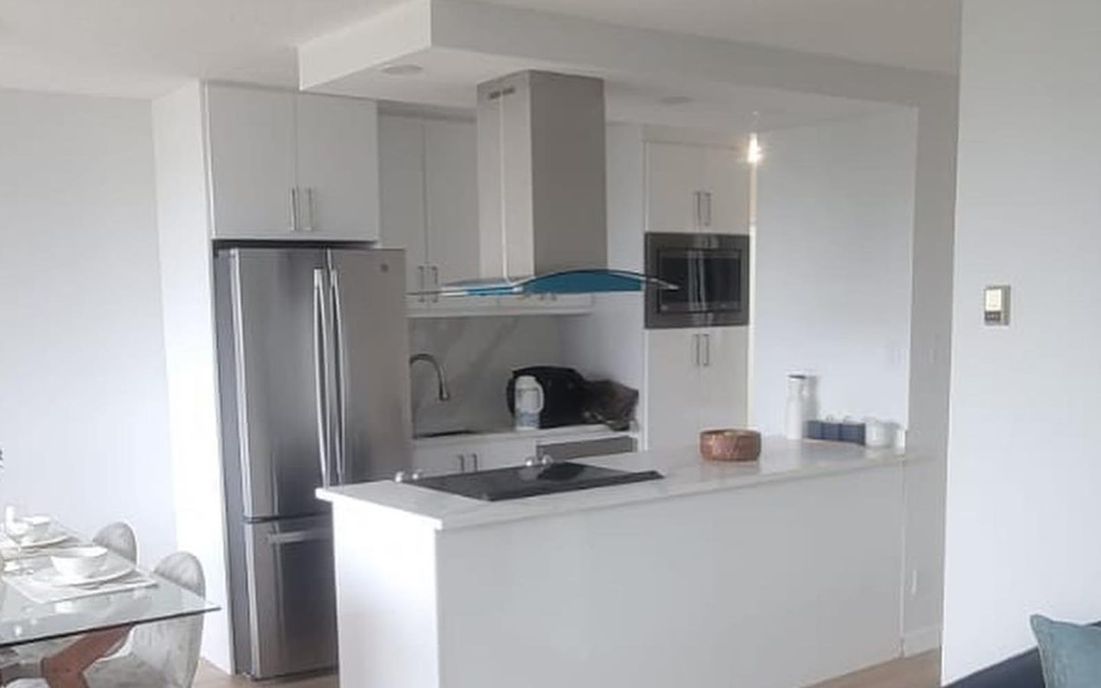 A completed apartment kitchen opened to the living area, with a white island, induction cooktop, chimney hood and integrated appliances.